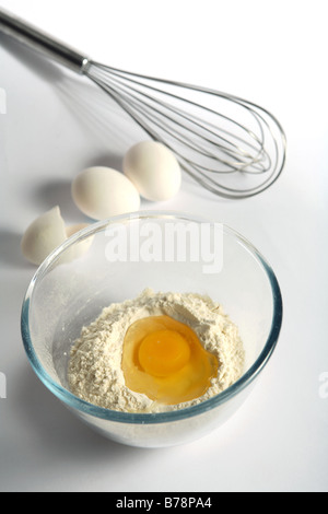 An egg broken into flour in a mixing bowl with other eggs and a whisk behind Stock Photo