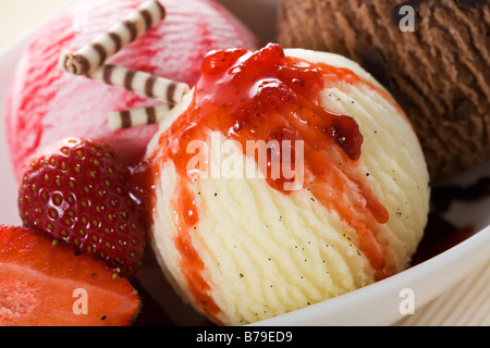 close-up of three balls of icecream - vanilla, strawberry, chocolate - with strawberry topping, strawberries and chocolate rolls Stock Photo