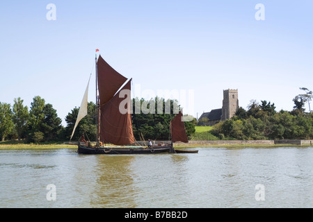 A Victorian spritsail barge glides under full sail past Iken Church on the River Alde in Suffolk UK Stock Photo