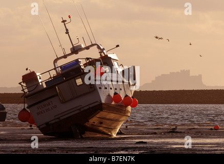 A fishing boat rests on its side in Holy Island harbour while the sun rises over Bamburgh Castle in the background Stock Photo