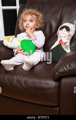 Two year old little girl wearing pajamas sitting in a big leather chair with her 2 handmade dolls Stock Photo