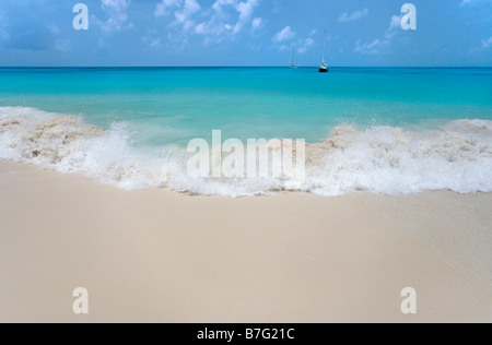 A wave breaks on 11 Mile Beach Barbuda West Indies as two cruising sailboats rest at anchor off shore Stock Photo