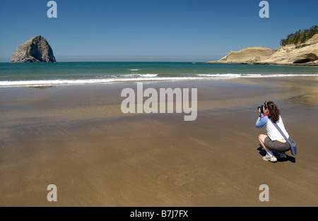 Woman (20-25) photgraphing on a sandy beach with blue sky and rock formations, Newport, Oregon Stock Photo