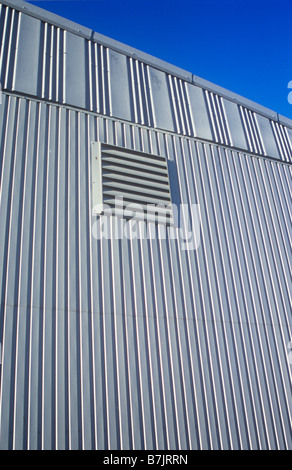 Detail in warm light of pale grey slatted wall and roof of warehouse or factory with large air vent under blue sky Stock Photo