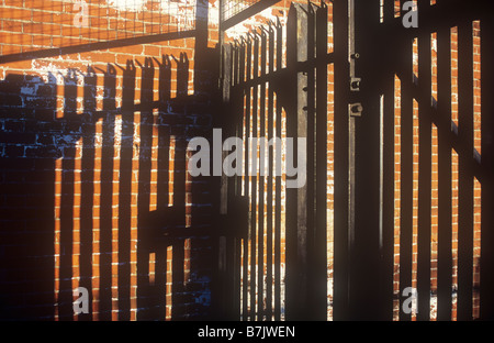 High black metal security gates topped with angled spikes and wire mesh casting deep shadows on orange brick wall in warm light Stock Photo