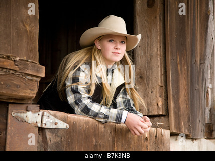 Teenage cowgirl leaning on stall door and looking out from old wooden stable on a farm Stock Photo
