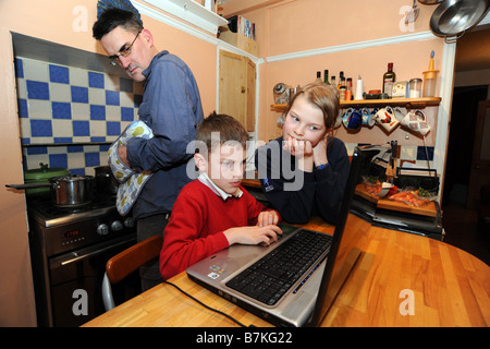 A father checks his children to see what they are doing on the laptop in the kitchen while he cooks tea. Stock Photo