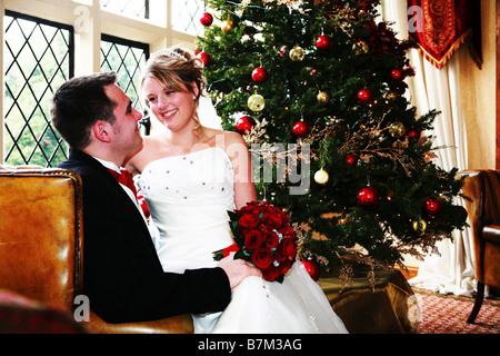 Bride and Groom at Xmas time winter wedding day cuddle in arm chair in front of Xmas Christmas tree warm and festive wedding day Stock Photo