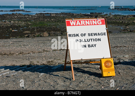 Sign at top of beach warning of sewage pollution in bay. In the distance is a slick at the waters edge caused by raw sewage. Stock Photo