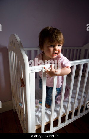 A unhappy looking  baby girl stands up in her cot . Stock Photo