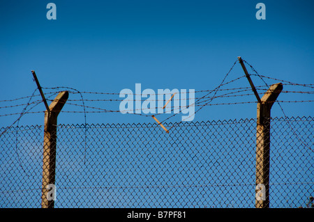 Concrete posts supporting a chain link fence topped with barbed wire, against a clear blue sky. Stock Photo