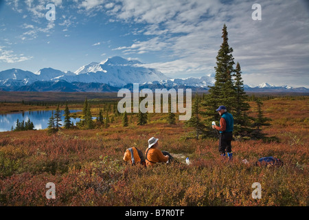 Two hikers rest on the tundra with the Alaska Range, Denali, and a small pond in the background in Denali National Park, Alaska Stock Photo