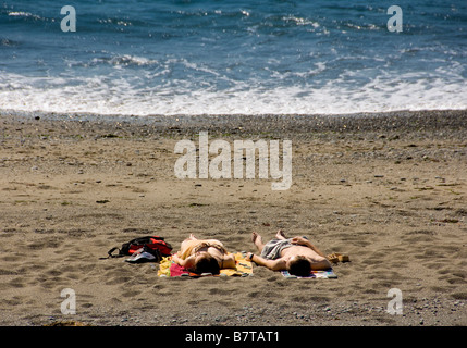 Unidentifiable caucasian couple laid on towels sunbathing on beach with sea in distance. UK Stock Photo
