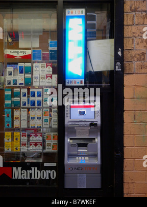 An outdoor automatic teller machine beneath a lighted 'ATM' sign next to a window display full of cigarettes in New York City. Stock Photo