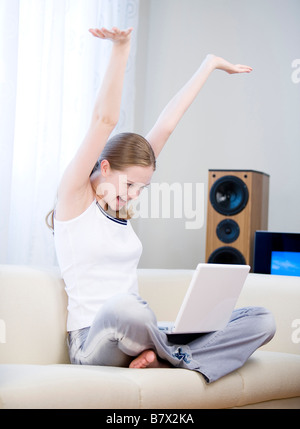 Young woman sitting on sofa cross legged with laptop stretching arms above head Stock Photo