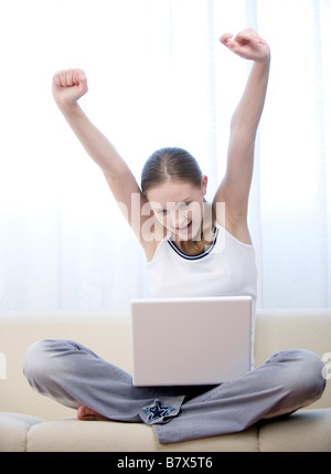 Young woman sitting on sofa cross legged with laptop stretching arms above head Stock Photo