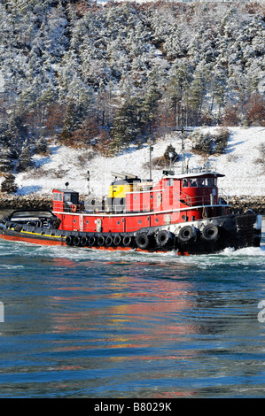 Old style red tugboat moving through water on a cold winter day with a hill of snow covered trees on shore Stock Photo