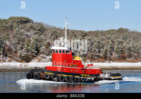 [Tractor tug]moving through the water on a cold winter day with snow covered trees on shoreline and exhaust showing from engine Stock Photo