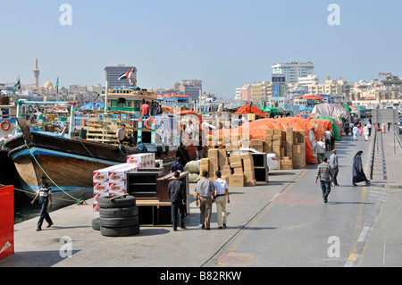 Dubai Creek worker on dhow boat & people walking past freight & merchandise on crowded congested port quayside United Arab Emirates UAE Middle East Stock Photo