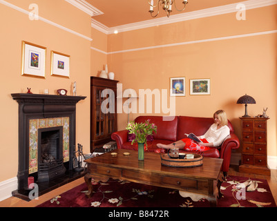 traditional living room with lady reading on red leather sofa with wooden coffee table and original cast iron fireplace Stock Photo