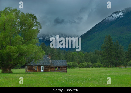 Old wooden house in a lush green field with mountains and a dramatic cloudy sky in the background Stock Photo