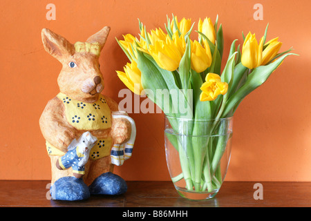 Fresh yellow tulips blossoming in a glass vase and paper mâché easter bunny in front of warm earthy colored wall Stock Photo