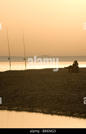 Fisherman  relaxing in beach chair watching 2 rods stuck in sand near waterline Stock Photo