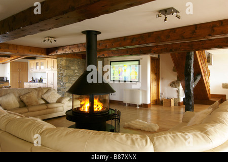 living room with large cream sofa and central circular lit fire with wooden beams and large television Stock Photo