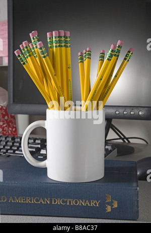 A cup or mug of number 2 Ticonderoga lead or graphite pencils and erasers in front of a computer and sitting on a dictionary Stock Photo