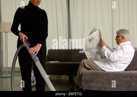 Woman is vacuum-cleaning while her husband is reading newspaper Stock Photo