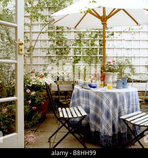 Breakfast table with blue and white checkered and fringed tablecloth and french iron cafe chairs in a London patio garden Stock Photo