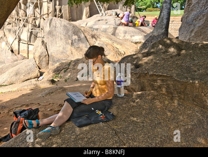 A female artist seated on a rock sketching ancient stone temple and other female workers cleaning stone monuments and gardens. Stock Photo