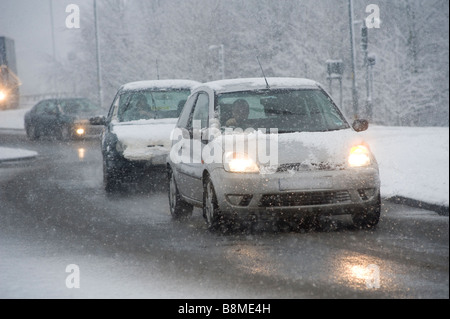 Cars driving slowly on a snowy road in winter in England with headlights covered in snow Stock Photo