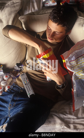 slobbish man lounging on a settee watching television and eating junk food Stock Photo