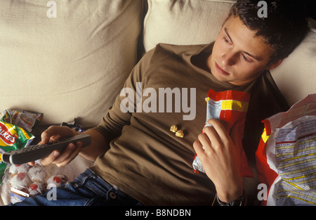 slobbish man lounging on a settee watching television and eating junk food Stock Photo