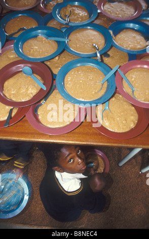 A young boy peers out from under a wooden table laden with blue and maroon bowls of porridge stacked on top of each other Stock Photo