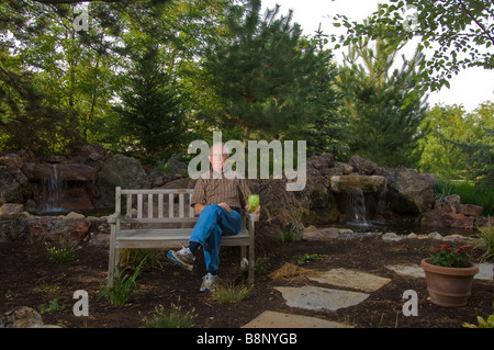 Senior man sitting on a bench by a man made pond and waterfall Stock Photo