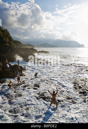 Tourists holding on to the rocky shore as a surprise big wave completely washes over the Queens bath near Princeville, Kauai, HA Stock Photo