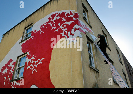 Graffiti artist on top of ladder painting wall mural of wave 'Stokes Croft' area of Bristol Stock Photo