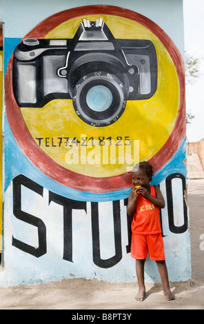 Little girl eating a piece of  mango in the street of Toubacouta in front of photographic studio wall painted sign  in Senegal West Africa Stock Photo