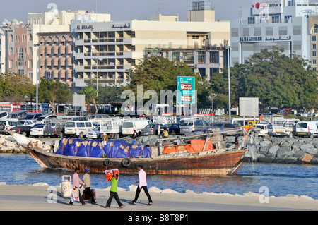 People & transport in Dubai Creek and fully loaded freight cargo on dhow boat on the move riverside car park beyond United Arab Emirates UAE Stock Photo