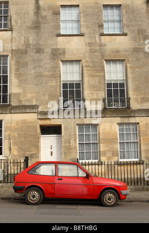 An old red tatty cheap car parked outside an expensive luxury Georgian terrace house in Bath England Stock Photo