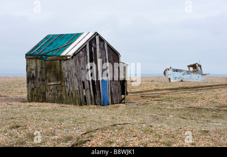 Disused wooden hut shack and shed and distant abandoned fishing boat on the shingle at Dungeness in Kent Stock Photo