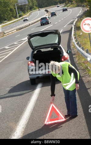 Woman wearing hi-viz placing a red warning triangle behind her broken down car on autoroute motorway slip road in France, Europe Stock Photo