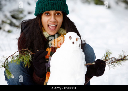 A happy, smiling woman wearing a hat and scarf with a snowman outdoors in the forest and snow. Stock Photo