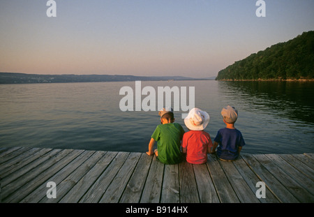 Three young boys sit on a dock on the Hudson River, New York,  at sunset Stock Photo