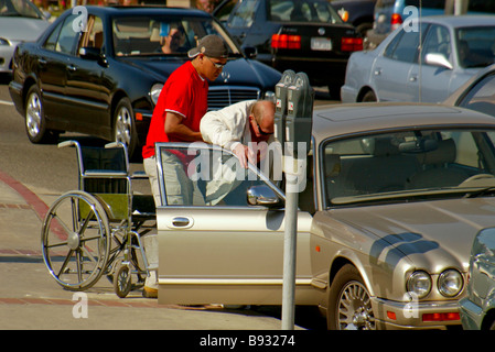 An African American professional caregiver helps an elderly handicapped man enter his car in Laguna Beach CA Note wheelchair Stock Photo