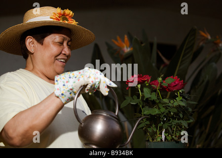 Senior Asian woman wearing straw hat outdoors watering flowers Stock Photo