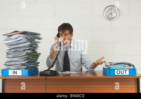 Stressed Office Worker working late into the evening with a large pile of work in his in tray. Stock Photo