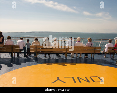 A group of people sit on the benches on the rear viewing platform of the 'Oscar Wilde' car ferry as it sails to Roscoff in sun Stock Photo